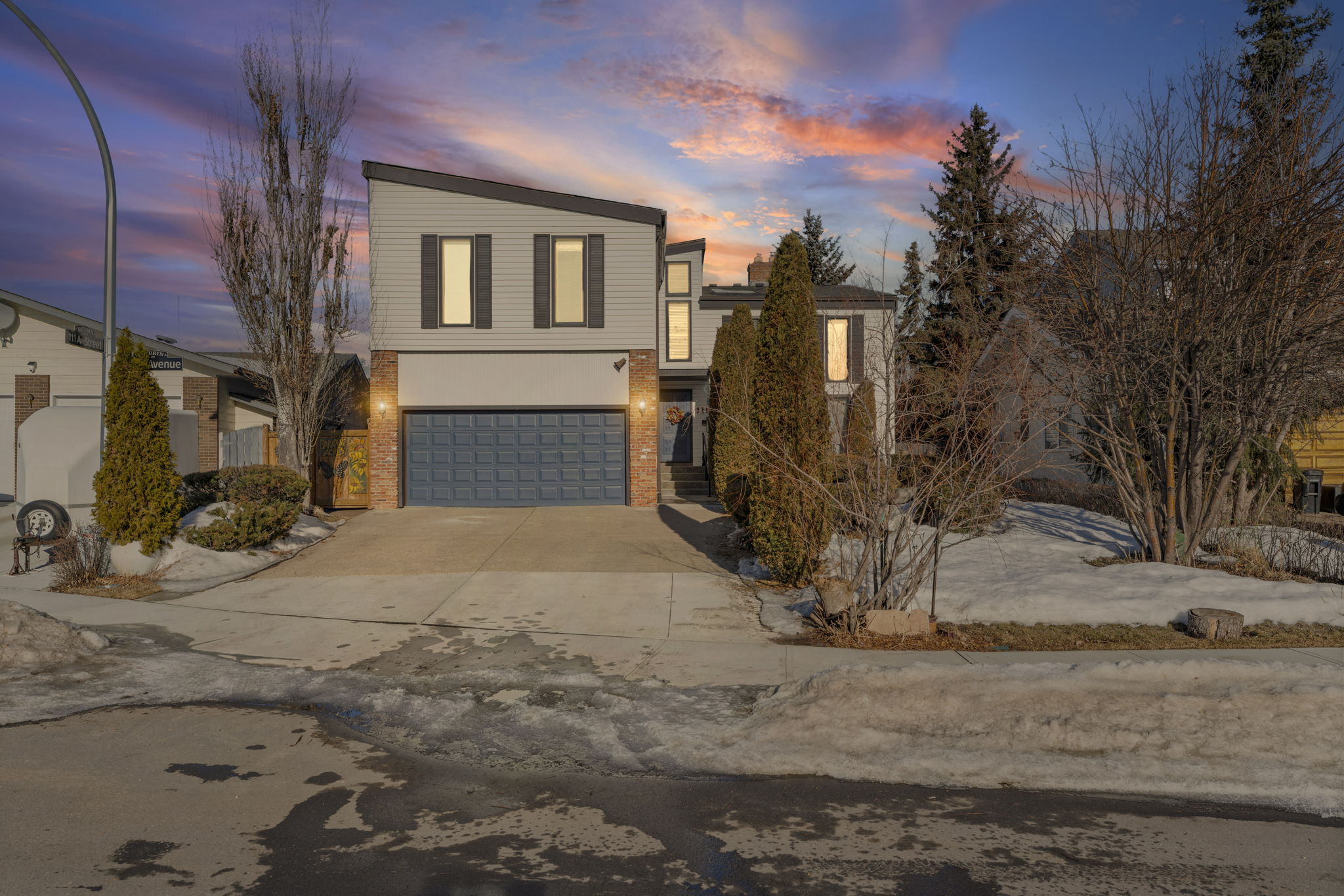 Front view of the 173a Avenue home at dusk with a blue garage door and snow along the driveway.