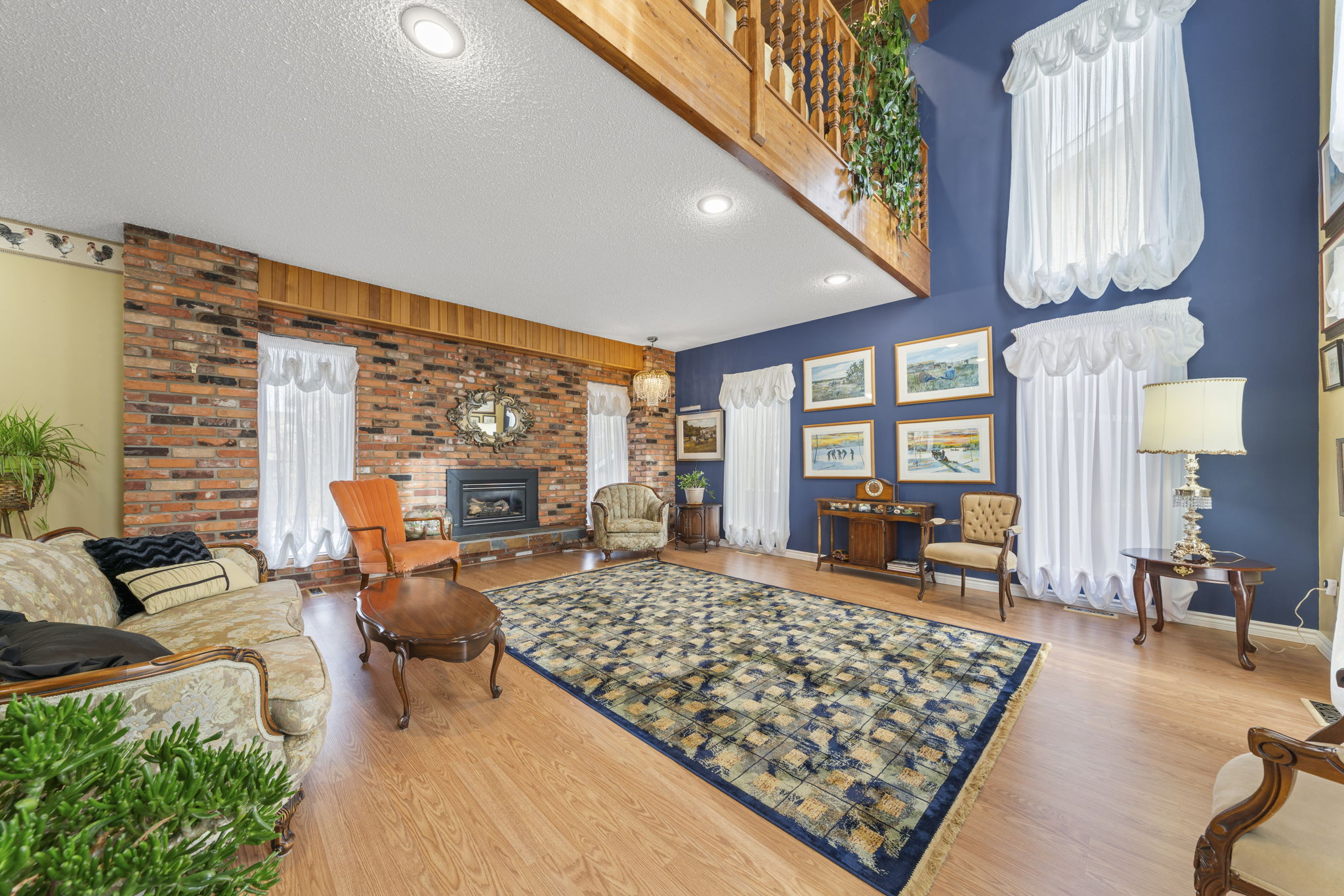 Double-height living room with a brick fireplace, patterned rug and wood balcony railing.