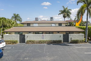 Front exterior elevation of the low-rise condo building showcasing the deeded parking area, shingle roof, and mature palm trees.