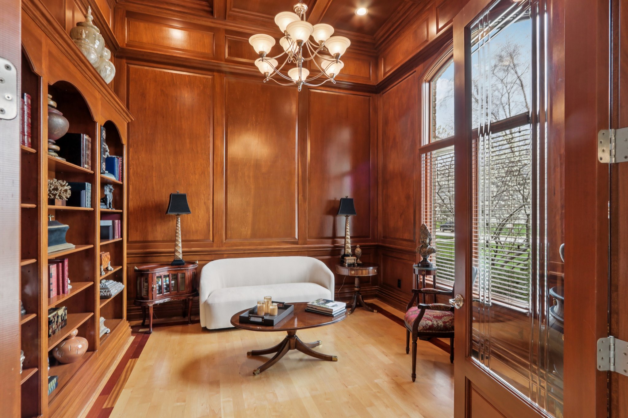 Elegant den/library boasts chandelier, floor-to-ceiling windows, built-in shelving, hardwood flooring and stately wooden walls with glass French doors.