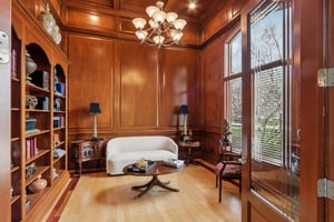 Elegant den/library boasts chandelier, floor-to-ceiling windows, built-in shelving, hardwood flooring and stately wooden walls with glass French doors.