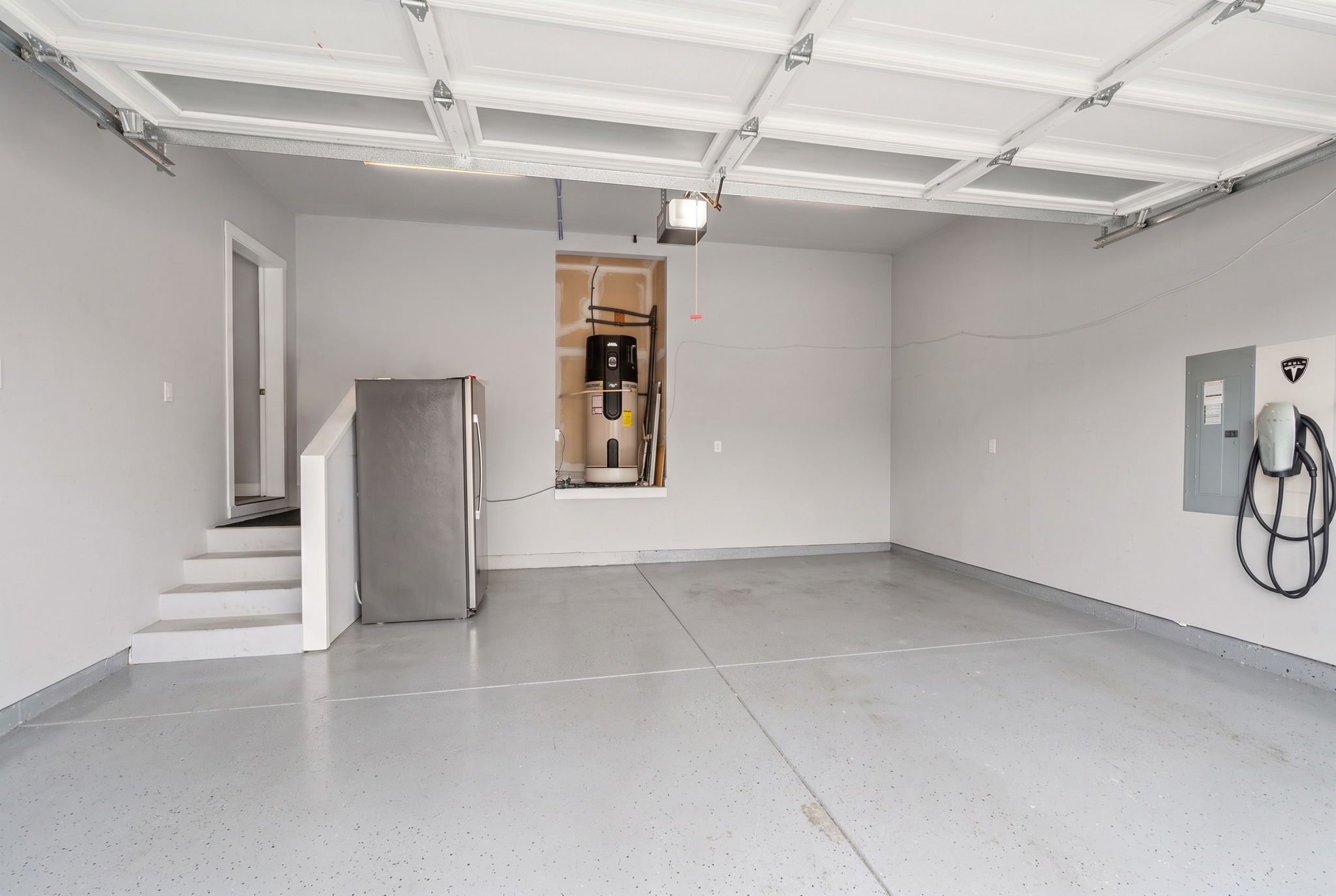 Spacious attached 2-car garage featuring tall ceilings, epoxy-coated floors, Tesla EV charger, and additional storage space.