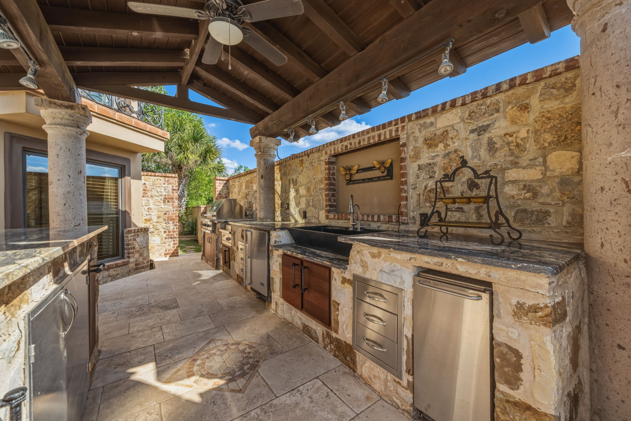 An expansive outdoor kitchen area featuring stone construction, granite countertops, and stainless steel appliances, all beneath a wooden beamed patio cover with a ceiling fan.  You can sit at the counter and enjoy your meal.