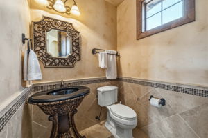 Half bath near kitchen features a decorative pedestal sink with a black basin, a toilet, a window with wood trim, and walls finished with tile wainscoting and a textured painted surface.