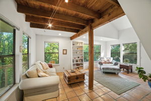 From the back door, the living room opens with walls of windows that frame the surrounding treetops, filling the space with natural light. Exposed wood beams and tile floors add rustic charm, while the indoor-outdoor flow creates a distinctive retreat feel not often found in the area.