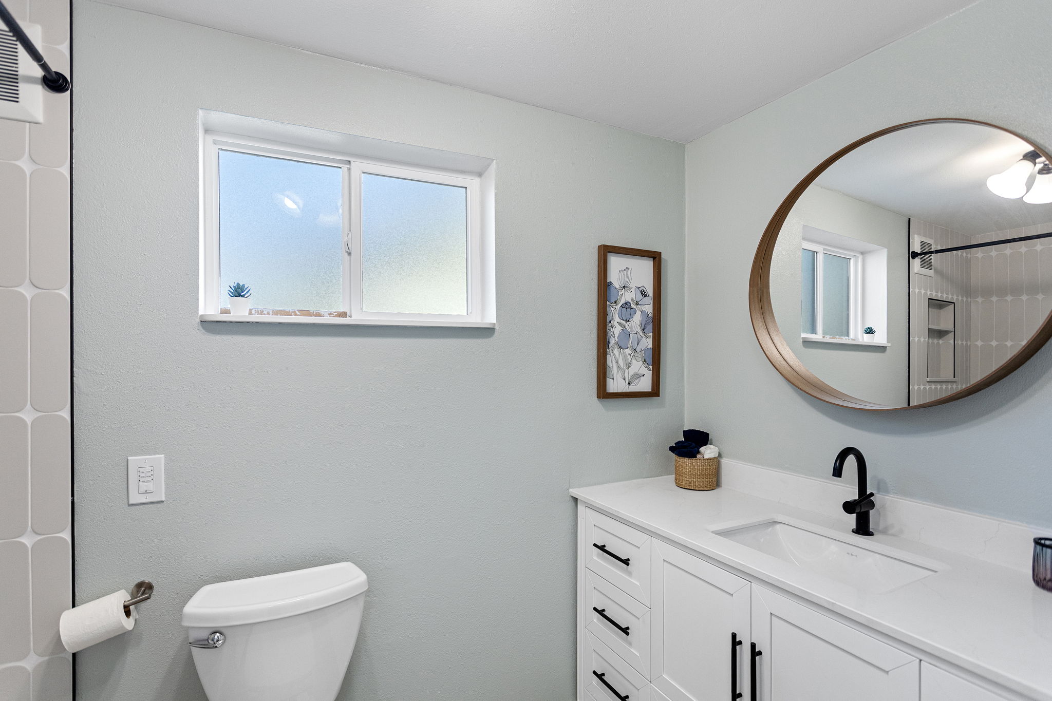 The full bath serves both daylight bedrooms — white shaker vanity, matte black hardware, warm wood-framed mirror, and a custom tile shower surround. Renovated, windowed, and finished with care.