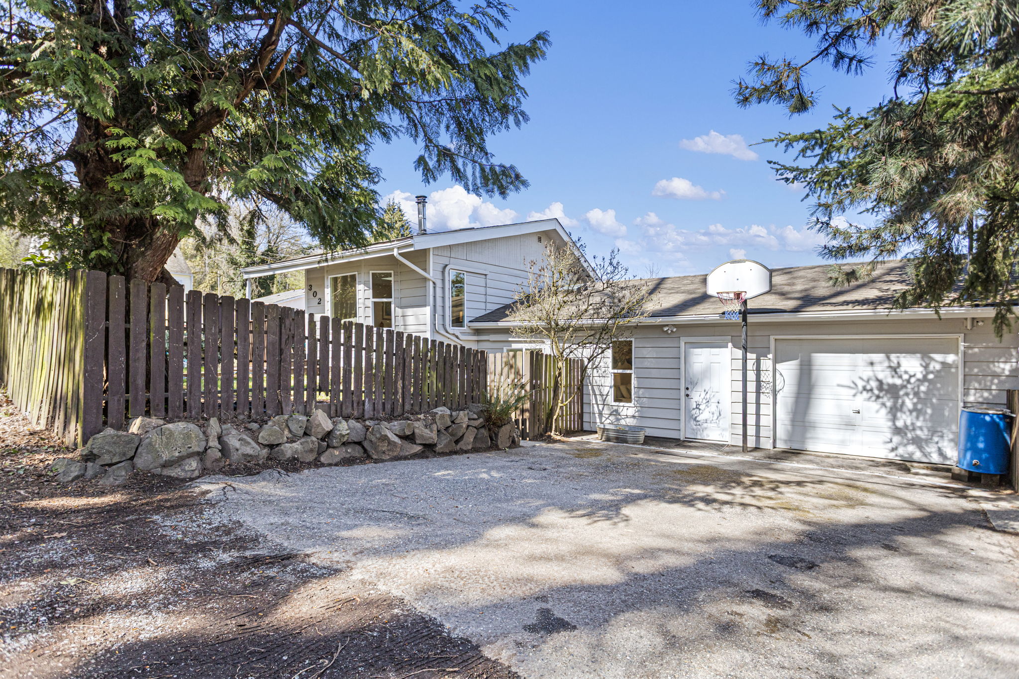 The driveway side: rock retaining wall, attached garage, and a basketball hoop that conveys. Mature trees on a fully fenced corner lot.