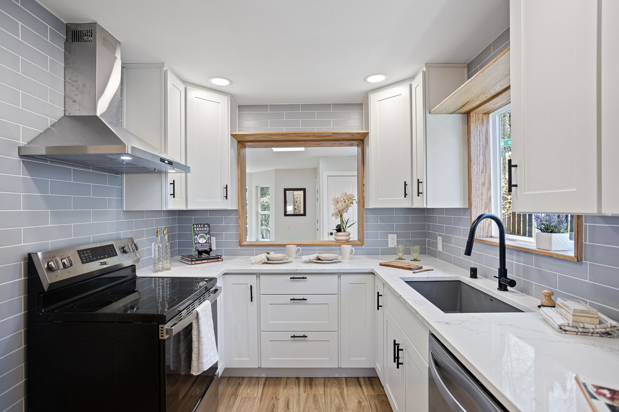 A kitchen reborn — new cabinetry, quartz countertops, blue-grey subway tile, stainless appliances, wall-mount range hood, and a wood-framed serve-through back to the living room.