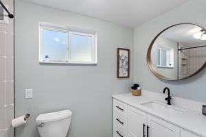 The full bath serves both daylight bedrooms — white shaker vanity, matte black hardware, warm wood-framed mirror, and a custom tile shower surround. Renovated, windowed, and finished with care.