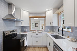 A kitchen reborn — new cabinetry, quartz countertops, blue-grey subway tile, stainless appliances, wall-mount range hood, and a wood-framed serve-through back to the living room.