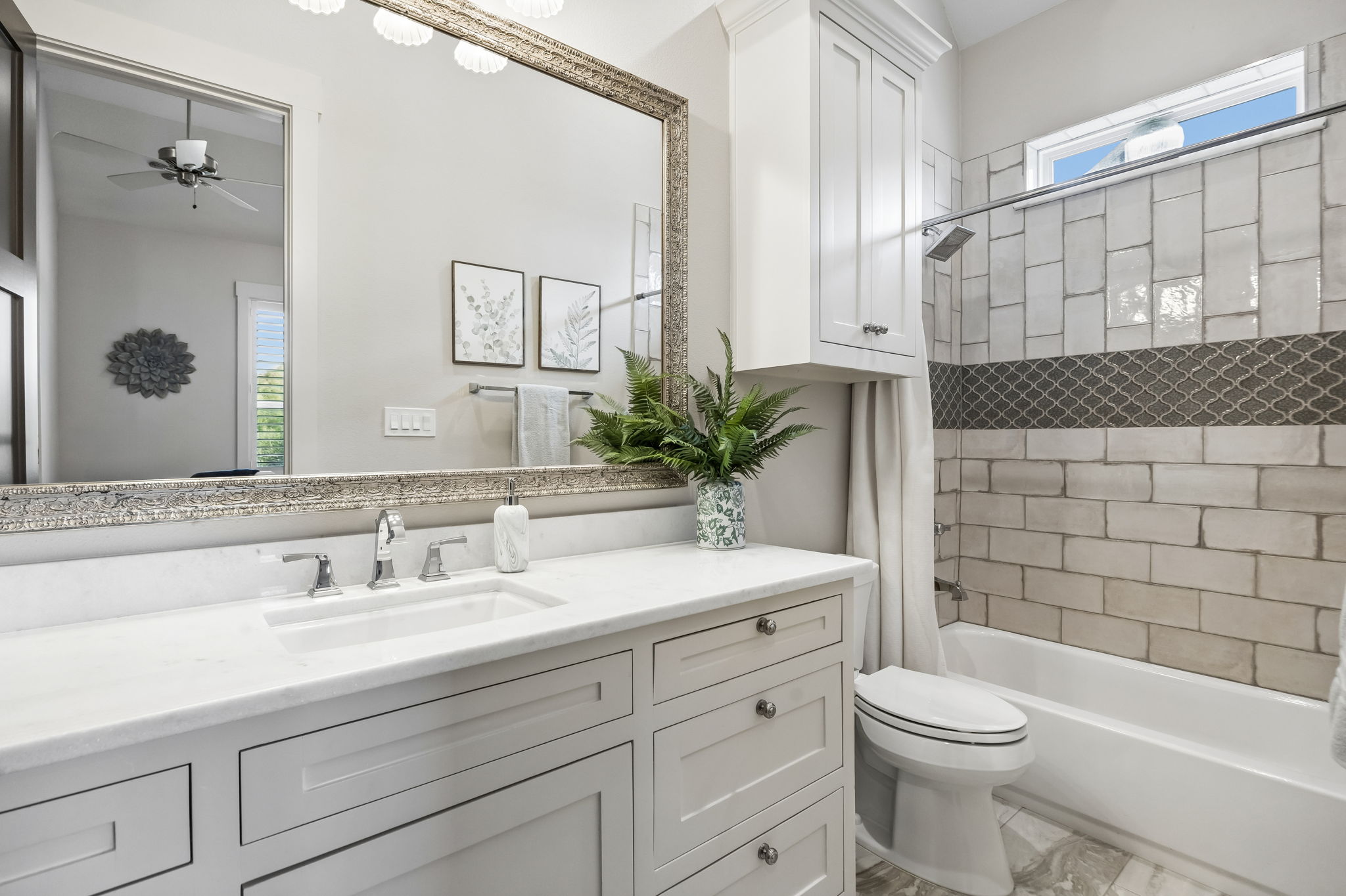 A Private Bath with Custom Tilework – The ensuite bath features a long quartz-topped vanity, abundant cabinetry, a tiled tub-shower surround, decorative accent detail, and a high window that brings in natural light.