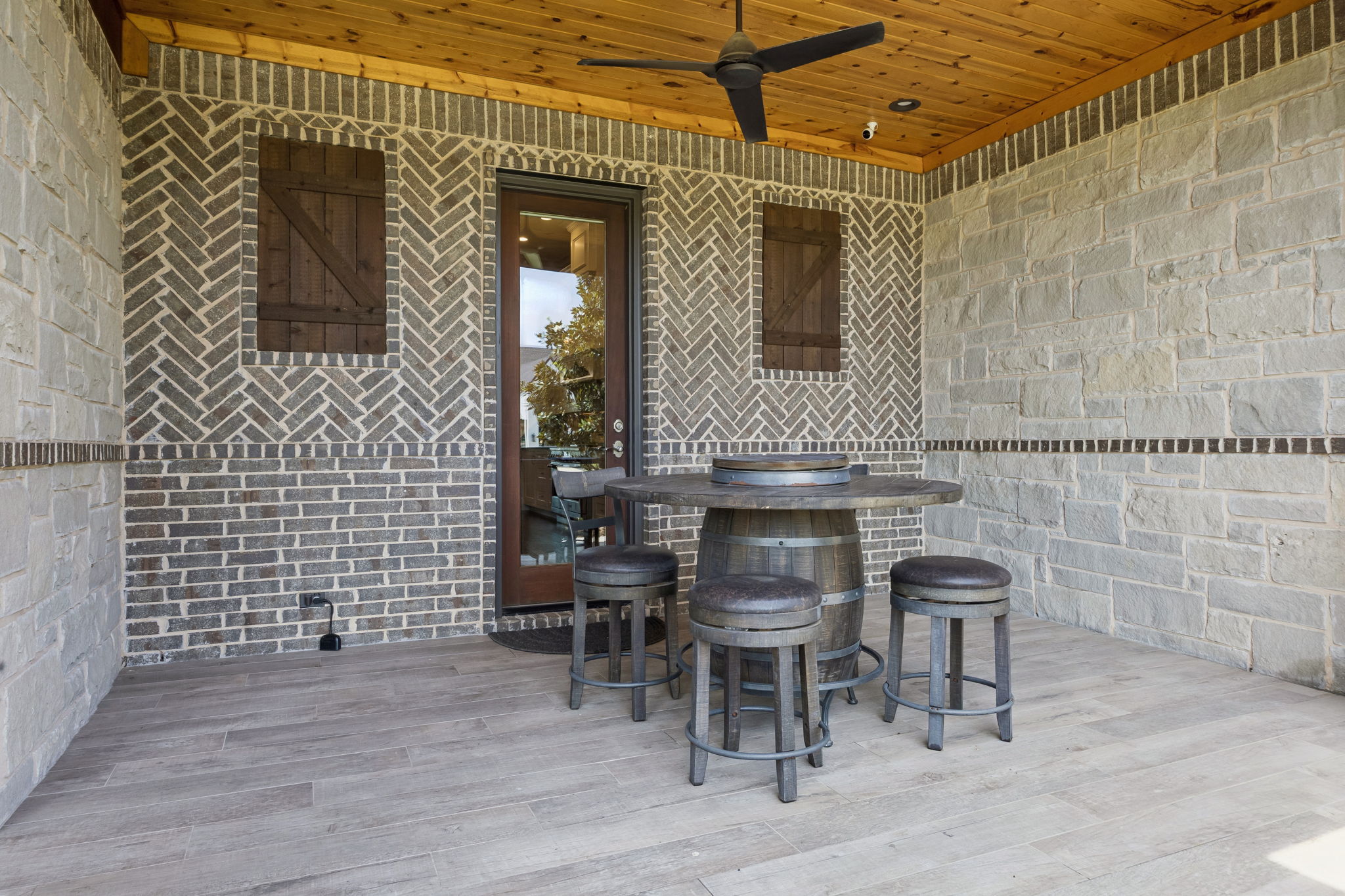 A Covered Side Patio with Custom Masonry – Herringbone brick, stone walls, a cedar ceiling, tile flooring, and a ceiling fan create a private outdoor room just off the interior living spaces.