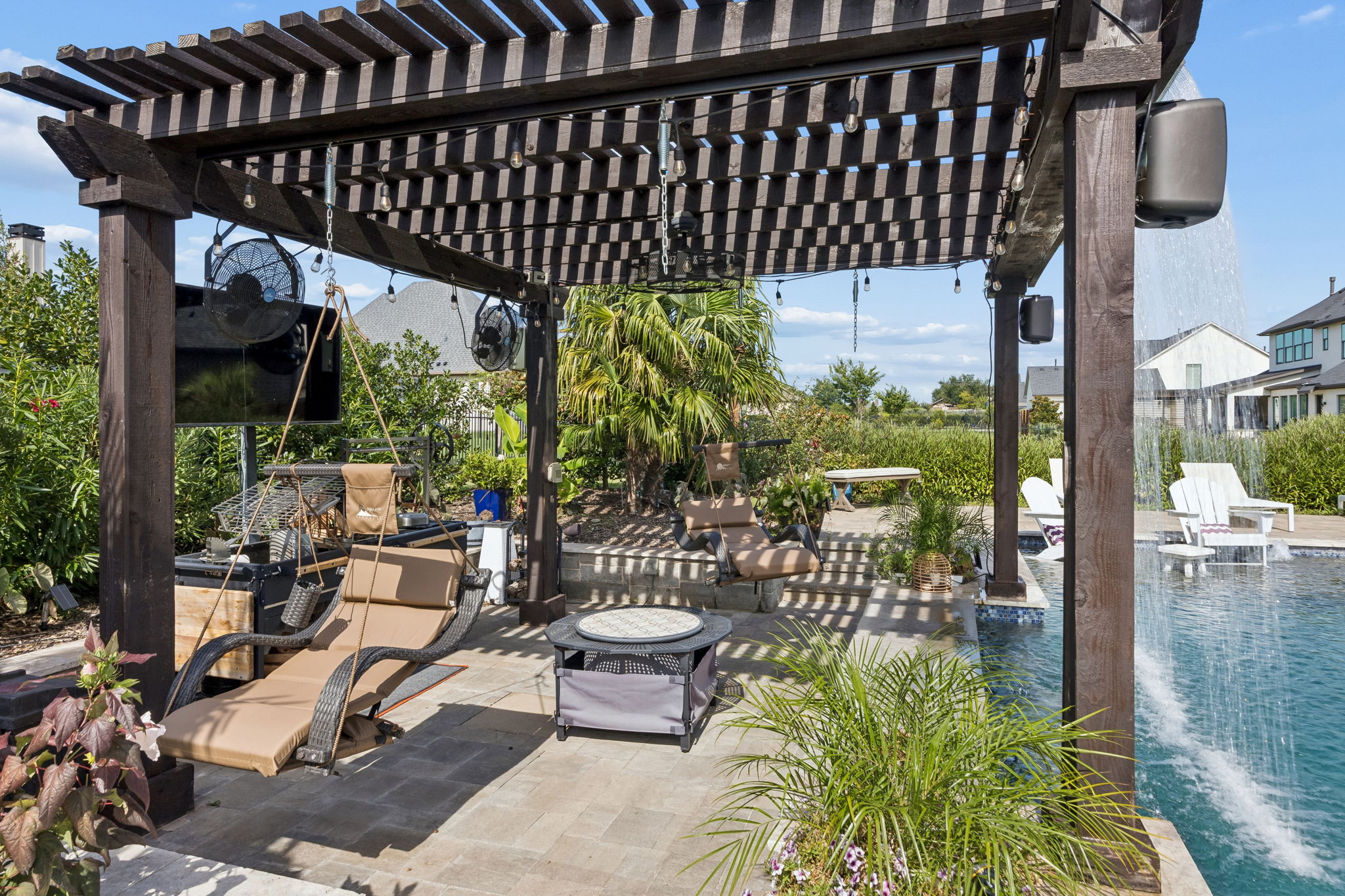 A Pergola Retreat Beside the Water – The cedar pergola adds another shaded destination with hanging seating, string lights, fans, and a dramatic curtain of water along the pool’s edge