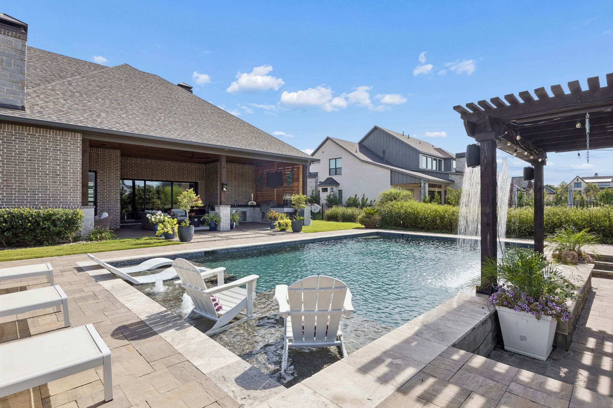 A Backyard View Worth Stepping Outside For – The pool stretches across the center of the yard, with the pergola, waterfall, shallow lounging area, and covered terrace creating a cohesive private retreat.