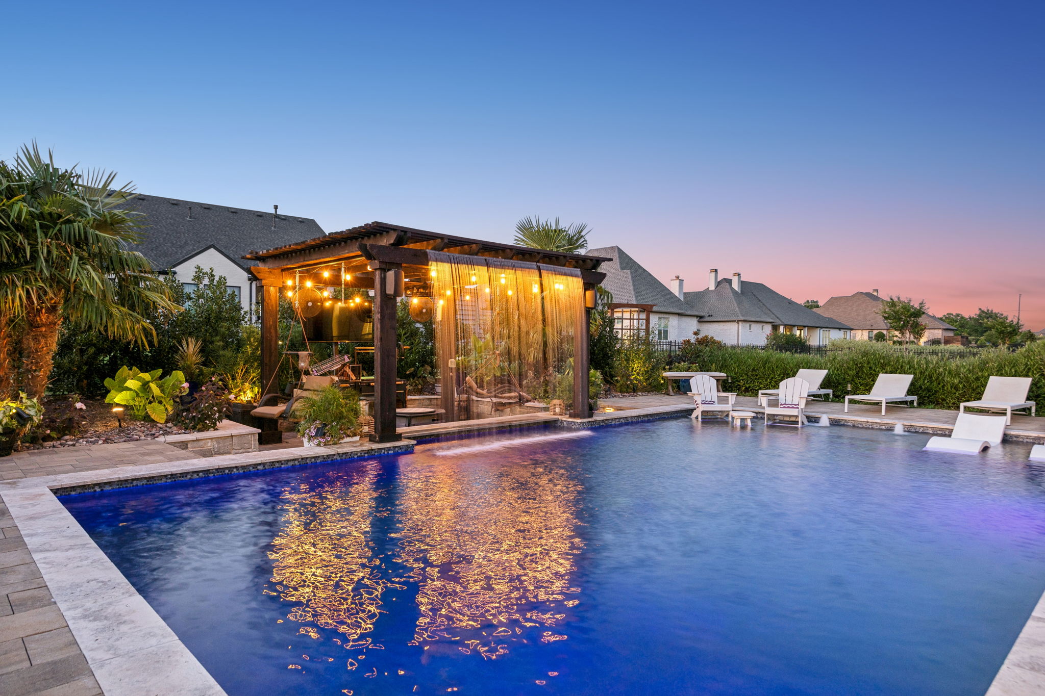 A Pergola That Becomes the Evening Focal Point – Warm string lighting, cascading water, and pool reflections turn this shaded retreat into a striking destination after sunset.