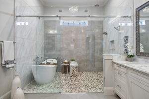 A Wet Room Designed for a True Spa Experience – The oversized glass enclosure brings together the freestanding soaking tub, dual shower stations, handheld fixtures, pebble flooring, and textured tile in one dramatic bathing space.