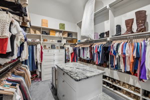 A Boutique-Style Primary Closet – A center storage island, floor-to-ceiling hanging systems, display shelving, and extensive shoe storage create a spacious dressing area designed around organization.