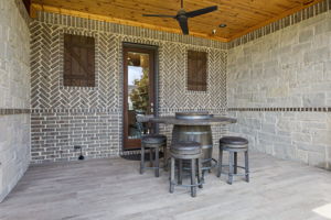 A Covered Side Patio with Custom Masonry – Herringbone brick, stone walls, a cedar ceiling, tile flooring, and a ceiling fan create a private outdoor room just off the interior living spaces.