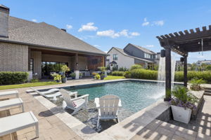 A Backyard View Worth Stepping Outside For – The pool stretches across the center of the yard, with the pergola, waterfall, shallow lounging area, and covered terrace creating a cohesive private retreat.