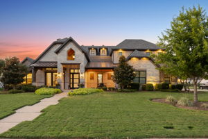 Distinctive Architecture at Sunset – Exterior lighting emphasizes the stonework, varied rooflines, metal accents, and covered entry as the evening sky adds warmth to the home’s curb appeal.
