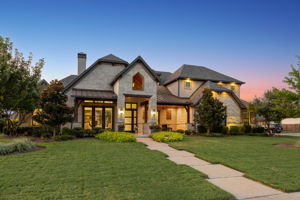 A Memorable Evening Arrival – Illuminated stone and brick details, a contemporary glass-paneled door, and layered rooflines give the home an inviting presence after sunset.