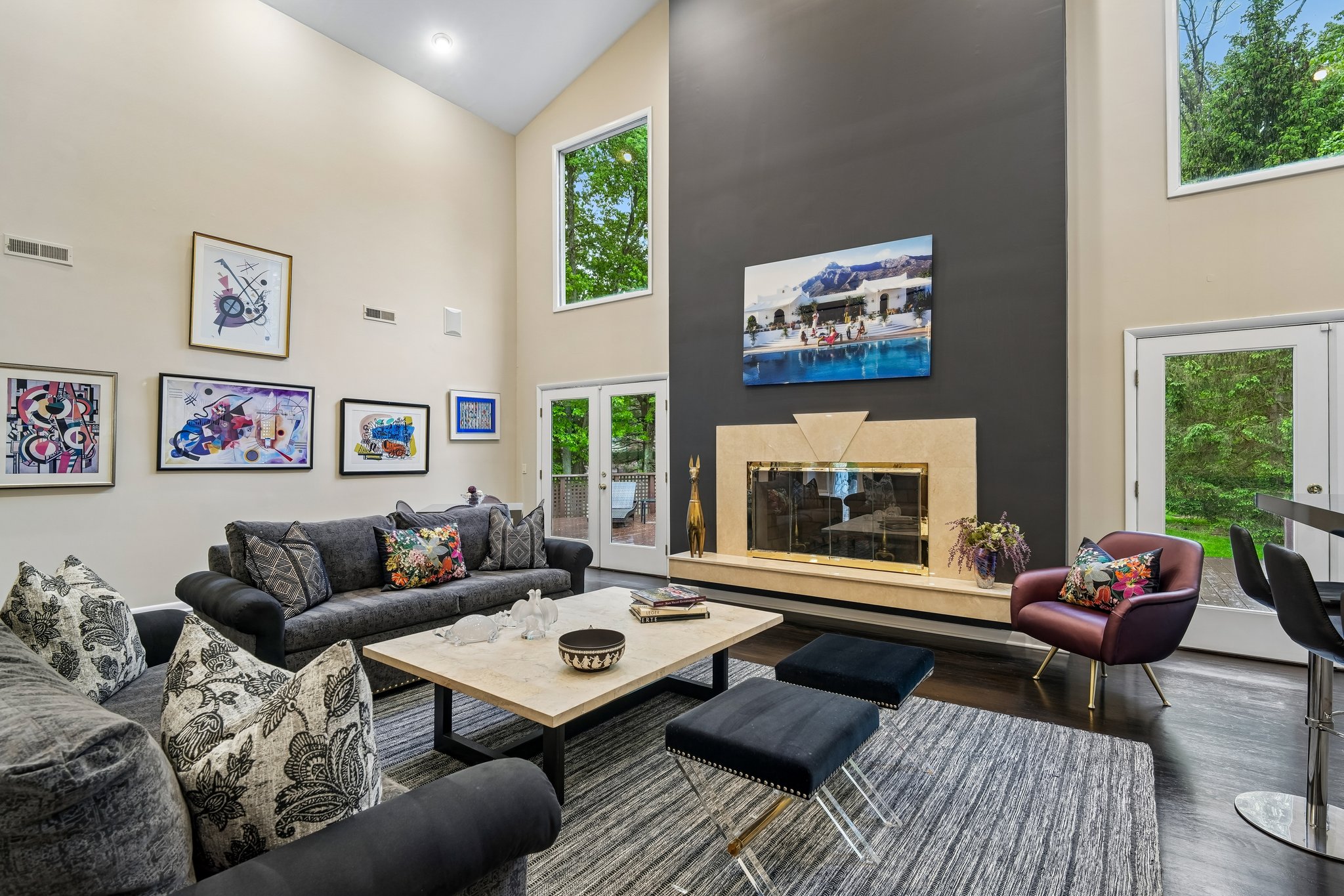 Experience unmatched scale in soaring ceiling living room, where stately fireplace commands attention. Sunlight pours in through expansive walls of windows/doors, creating an airy, light-filled sanctuary that seemlessly connects the interior warmth to the tranquil outdoor scenery.
