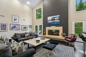 Experience unmatched scale in soaring ceiling living room, where stately fireplace commands attention. Sunlight pours in through expansive walls of windows/doors, creating an airy, light-filled sanctuary that seemlessly connects the interior warmth to the tranquil outdoor scenery.