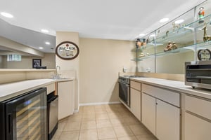 A finished lower level includes a convenient kitchenette that serves as the perfect refreshment station. With its own sink, cabinetry and fridge, the space is elevated by mirrored accent wall and glass shelving, offering both style and exceptional utility.