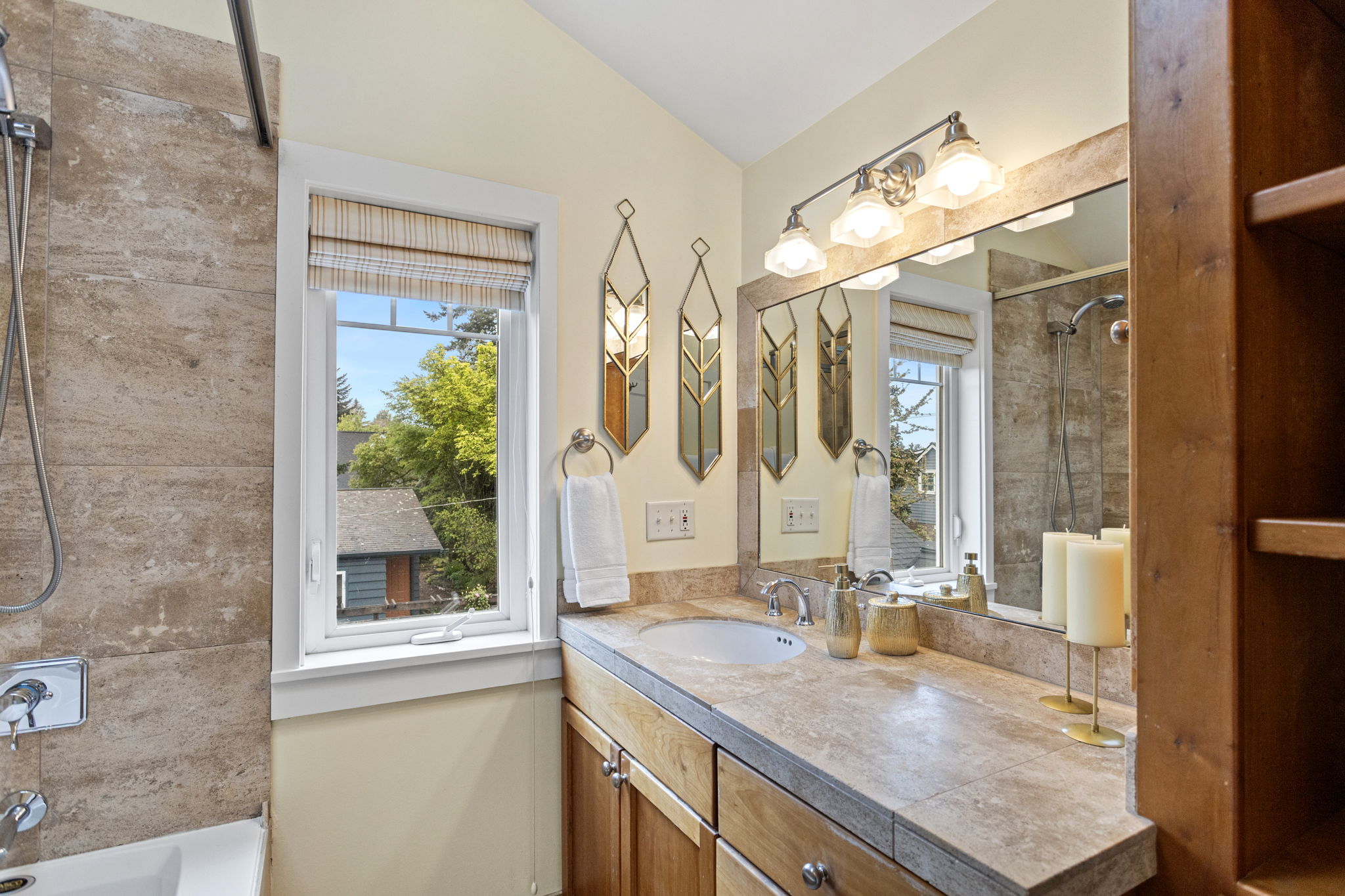Travertine tile, wood vanity, jetted tub, handheld shower, skylight overhead, built-in linen shelving. The primary bath doesn't ask for much — just blue sky and hot water.