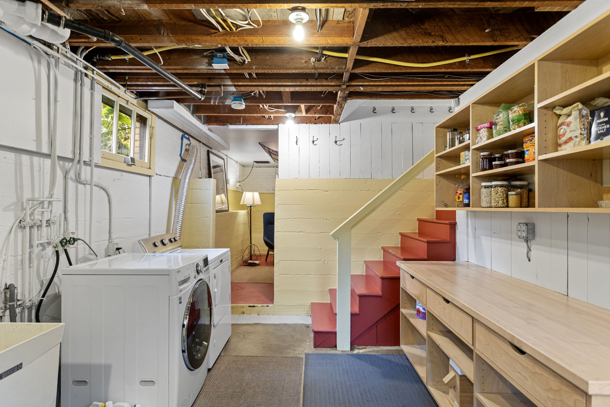 The laundry room does its job. Custom built-in shelving, full-size washer/dryer, utility sink. Down those red stairs: the bonus room.
