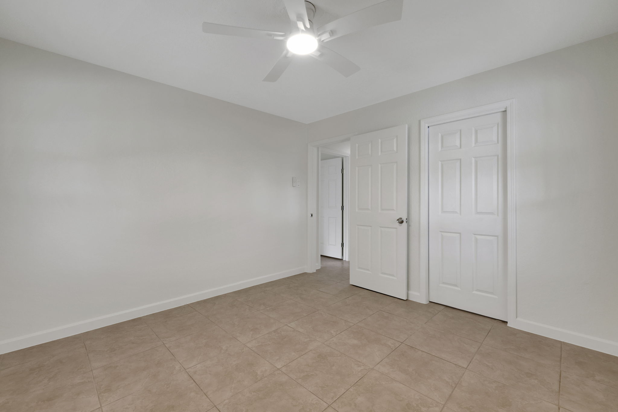 Primary bedroom features neutral-toned walls and tiled flooring, providing a clean and modern look. It includes a ceiling fan for comfort and two white doors, offering access and storage options. Ideal for a bedroom or office space.