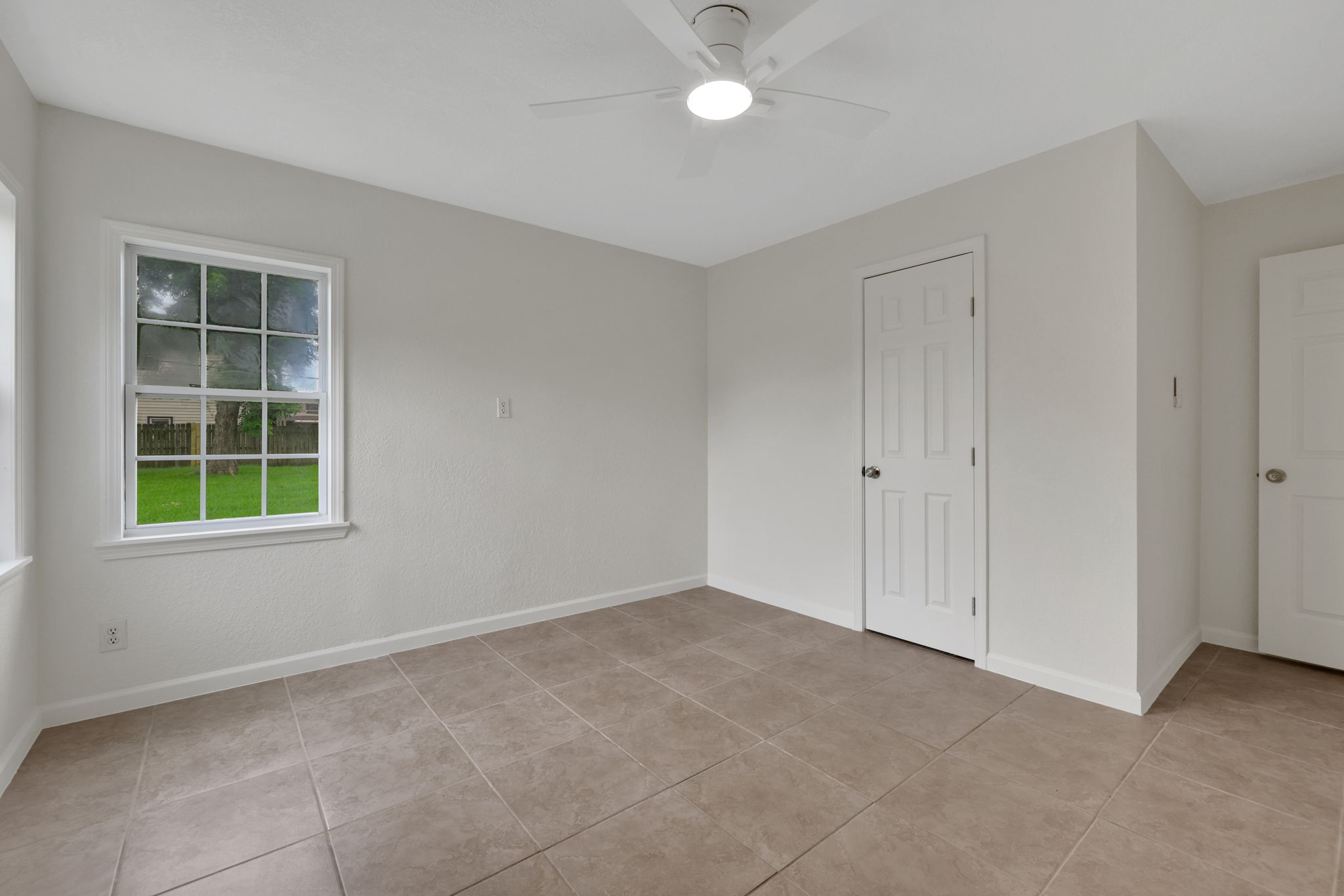 Bright, neutral-toned room with tiled flooring, a ceiling fan, and a large window offering a view of the green outdoors. Features two doors for access or storage. Ideal for a bedroom or office space.