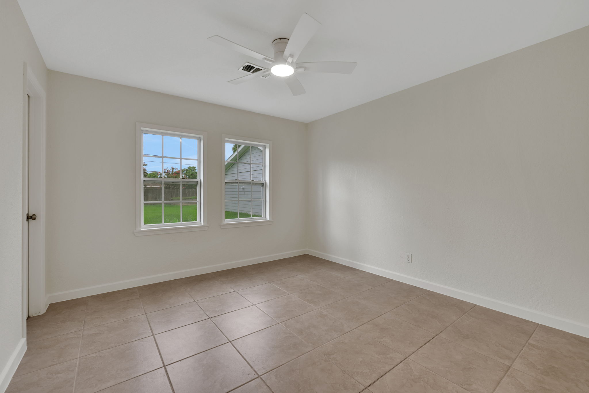 Bright, neutral-toned room with tiled flooring, ceiling fan, and two windows providing natural light. Ideal for a bedroom or office space.