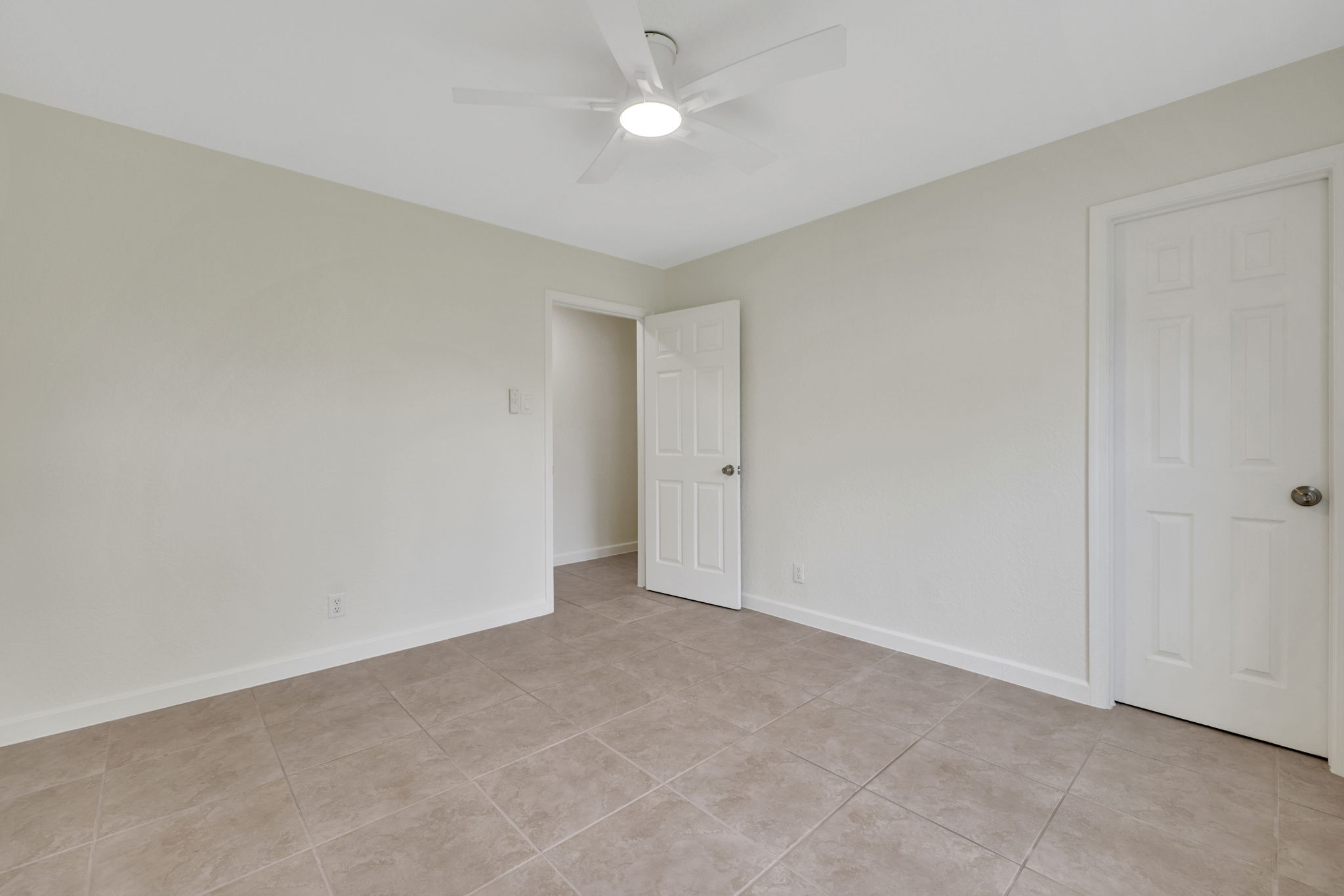 Secondary bedroom 1 with neutral-toned walls and tile flooring. It features a ceiling fan and two white doors, offering a clean and versatile space.