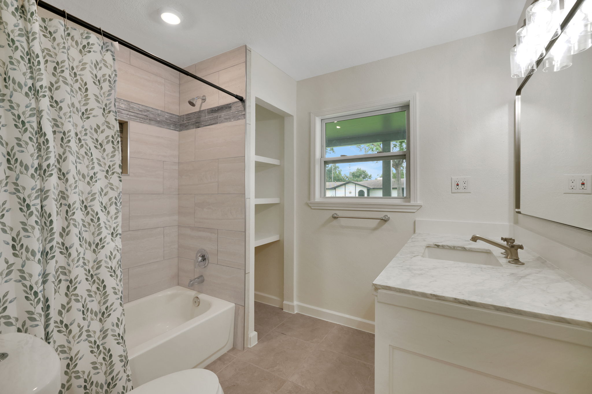Additional view of the bathroom features a clean and modern design with a vanity with a quartz countertop, a large mirror, a window providing natural light and built-in shelving for storage.