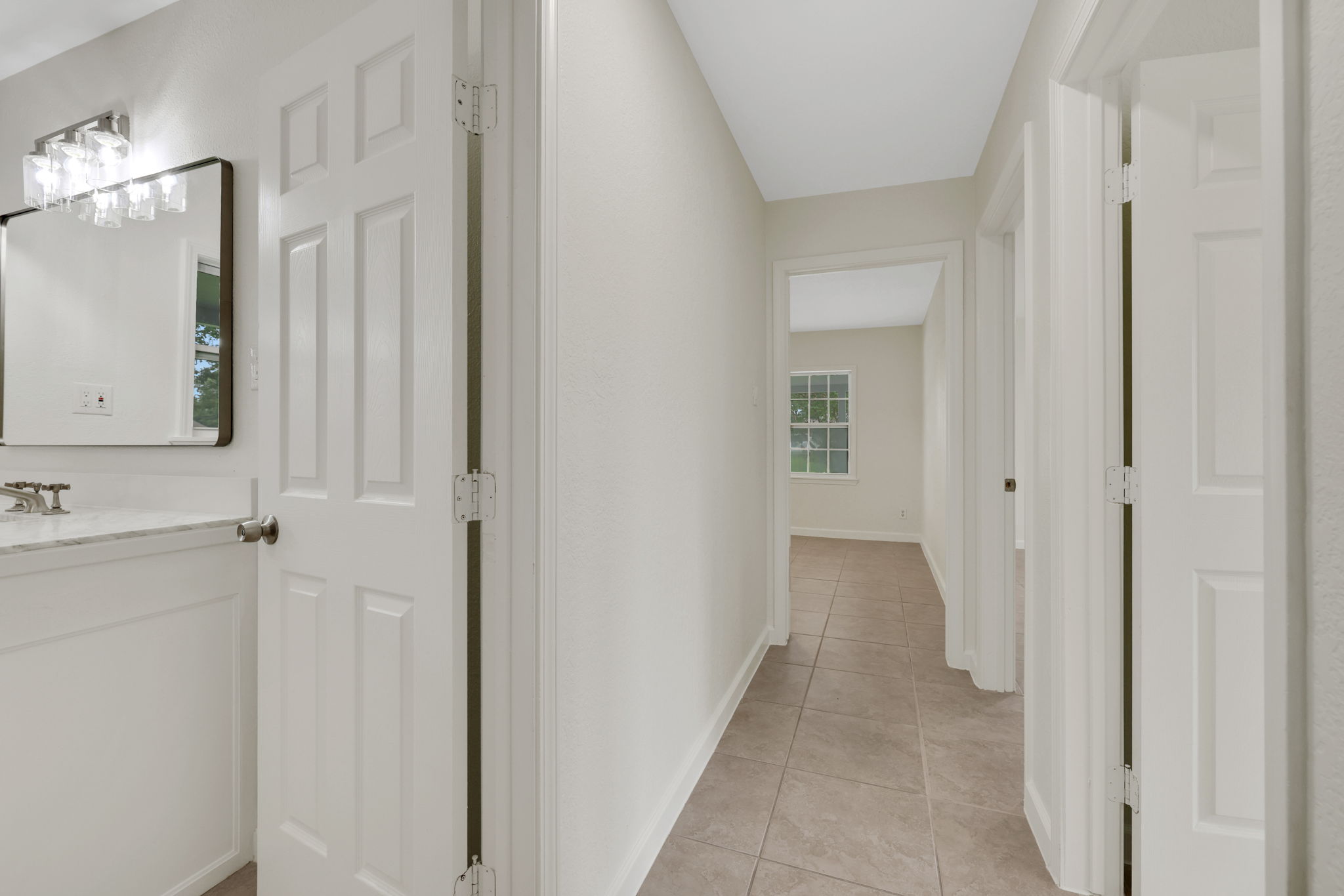 Bright, clean hallway with tiled flooring, leading to a room with a large window. A bathroom with a modern vanity and mirror is adjacent left.