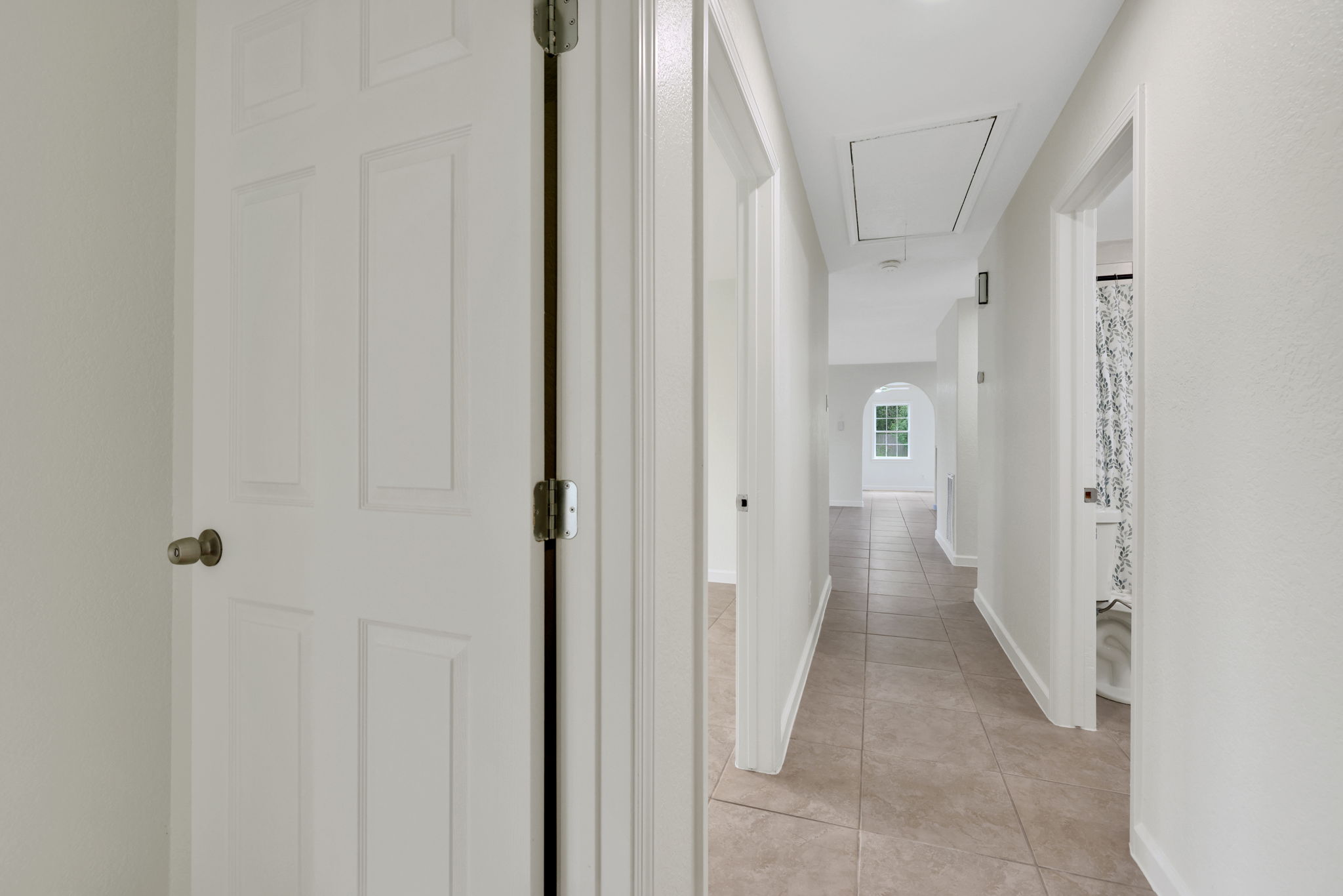 This hallway features light-colored walls and tiled flooring, leading to rooms on either side and an archway with a window at the end, creating a bright, open feel.
