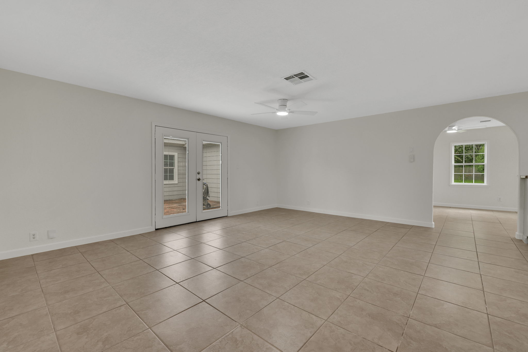 This spacious living area features neutral tile flooring, a ceiling fan, and French doors leading to an outdoor space. An arched doorway connects to another light-filled room, enhancing flow and openness.