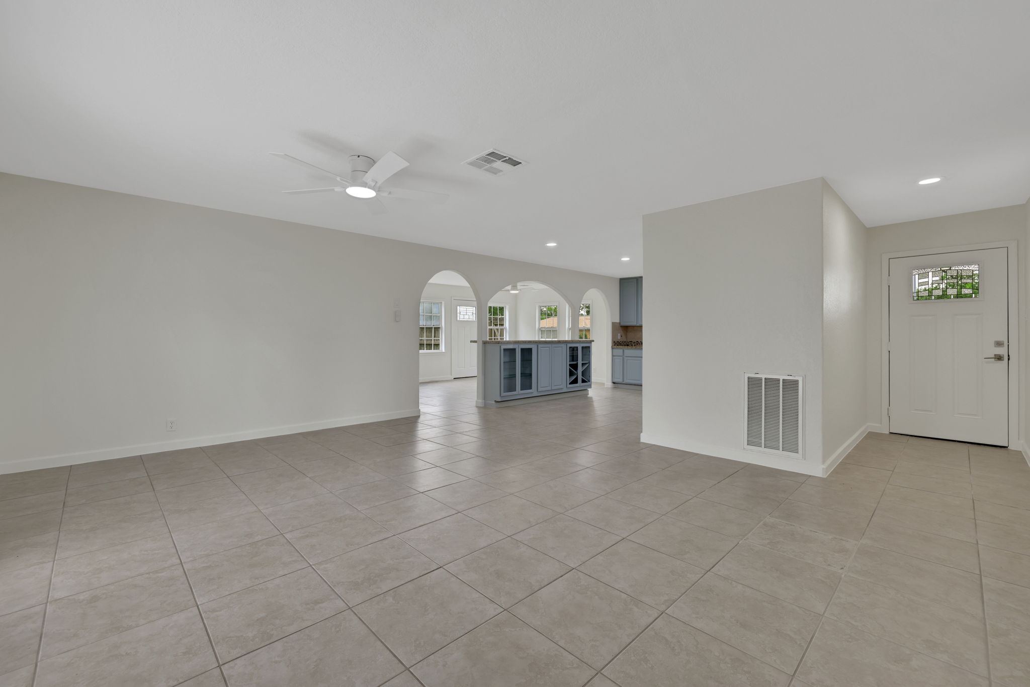 Spacious open-concept living area with tile flooring, neutral tone walls, and circulating air. Arched doorways lead to a modern kitchen with custom cabinetry and ample natural light.