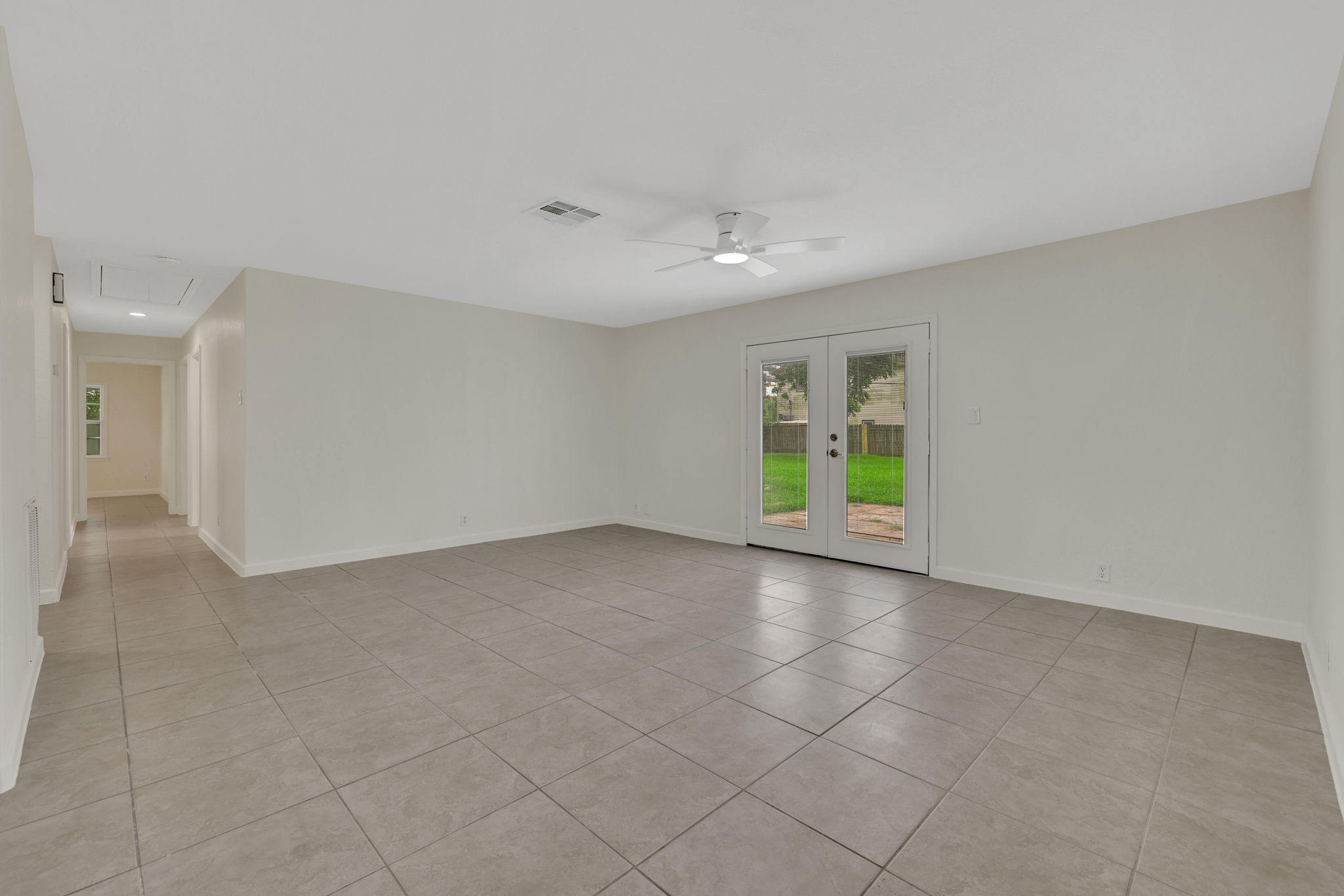 Spacious living area with neutral tiles, ceiling fan, and double doors leading to a plush backyard. Bright and open, perfect for family gatherings.