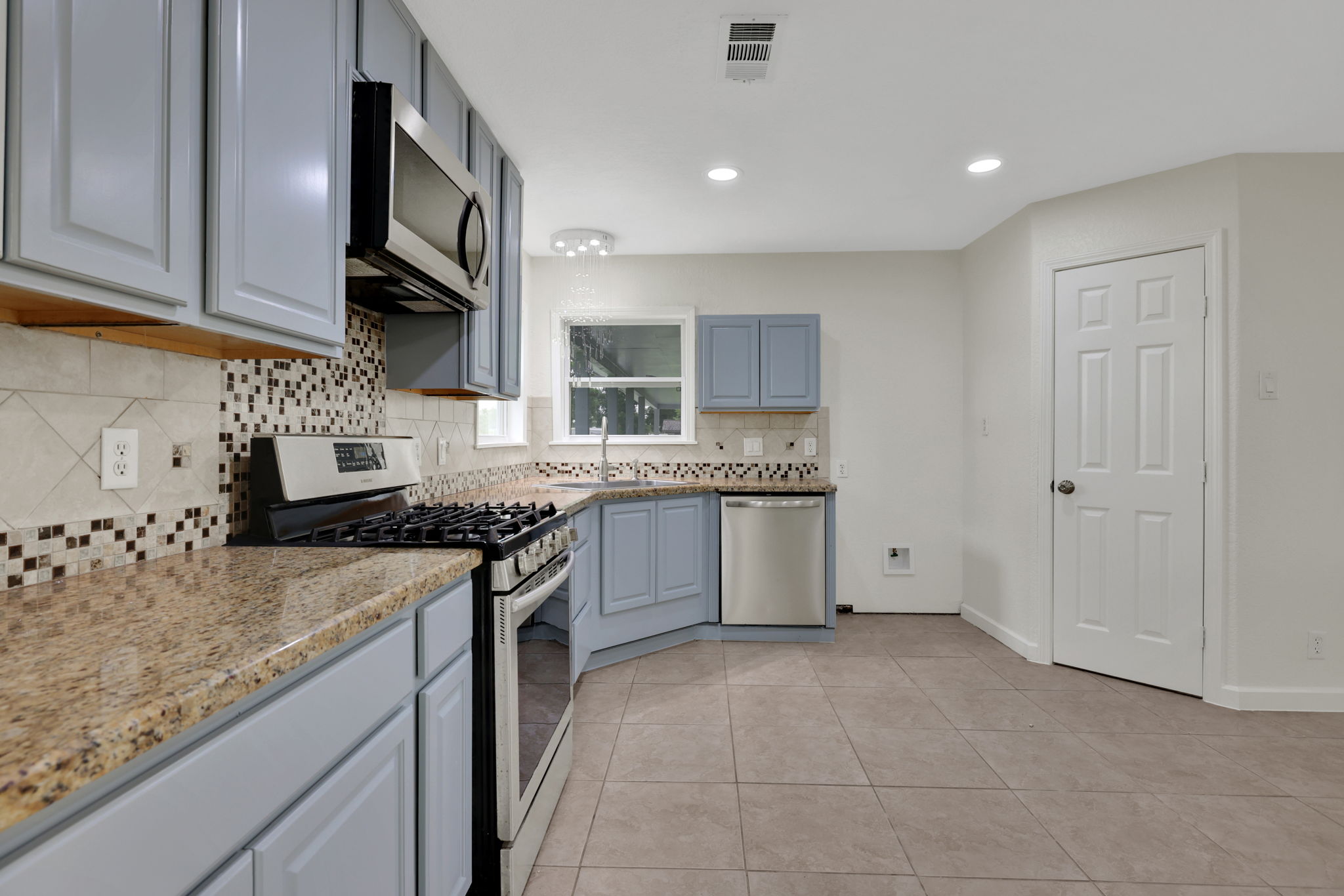 Modern kitchen with granite countertops, mosaic tile backsplash, and light teal cabinets. Features stainless steel appliances, including a gas stove and dishwasher, with ample natural light and tile flooring.