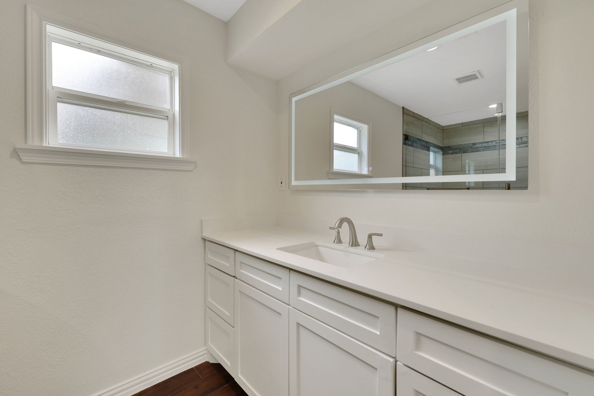 This modern bathroom features a sleek white vanity with ample storage, a large illuminated mirror, and natural light from a frosted window, creating a bright and airy atmosphere.
