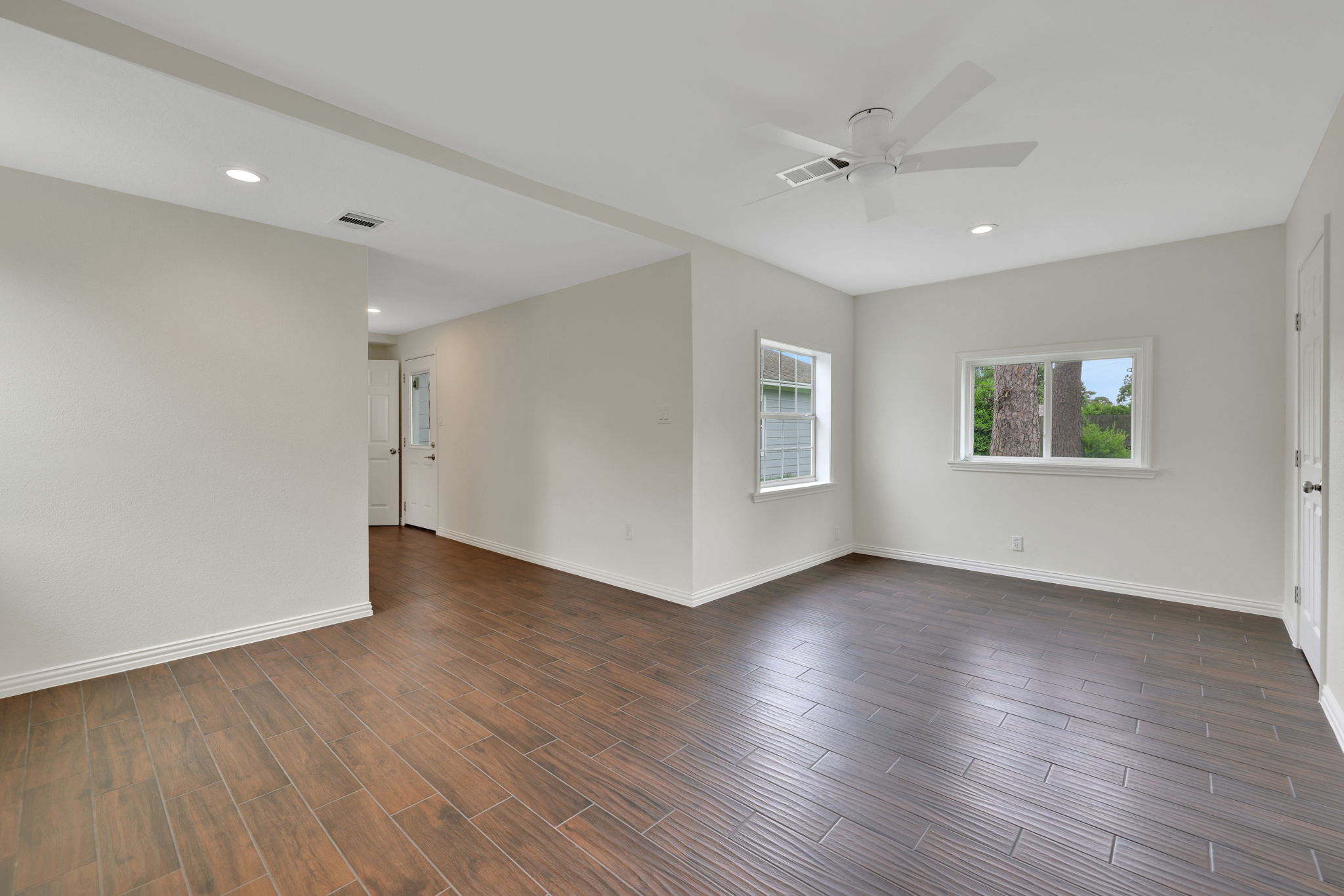 Bright and spacious room with sleek wood-look flooring, neutral walls, recessed lighting, and a ceiling fan. Two windows provide ample natural light, and there's a door leading to another area of the home.