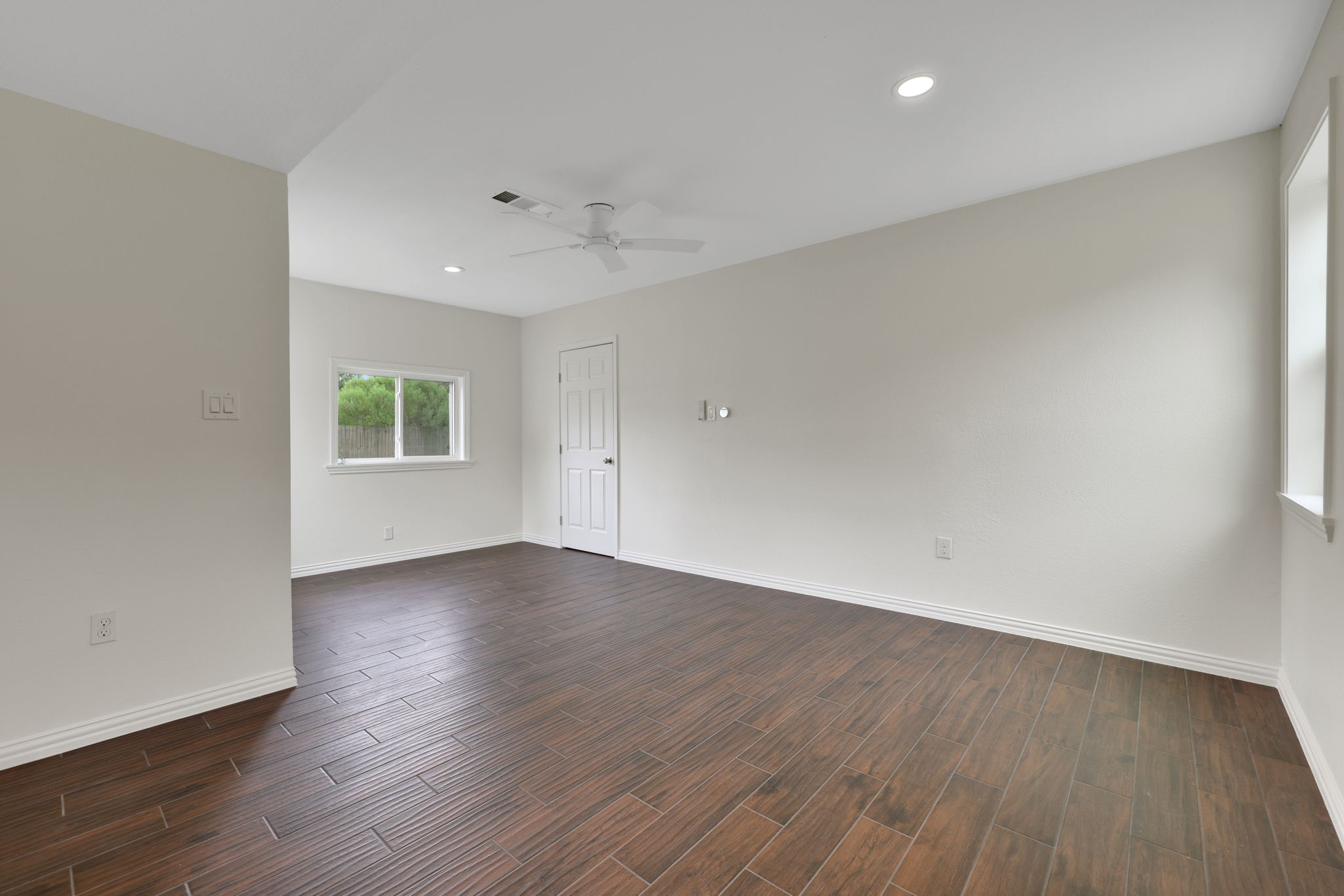 This room features sleek wood-look tile flooring, neutral walls, and ample natural light from multiple windows. It includes a ceiling fan and recessed lighting, providing a modern and airy feel.