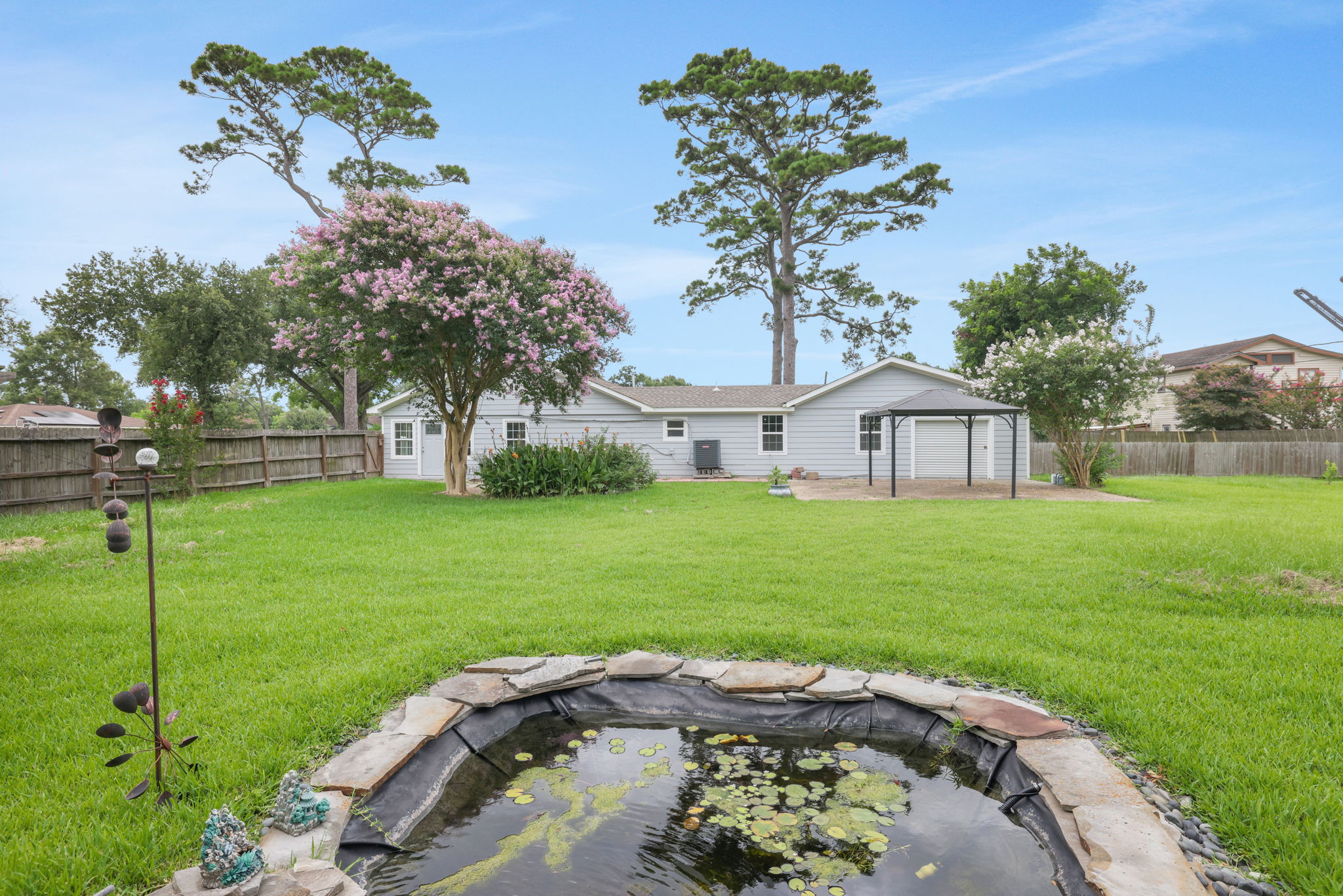 Spacious backyard with lush green grass, a small pond, and mature trees. The view includes a view of a fenced perimeter, and two patios, offering a serene outdoor space for relaxation or entertaining.