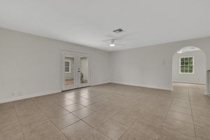 This spacious living area features neutral tile flooring, a ceiling fan, and French doors leading to an outdoor space. An arched doorway connects to another light-filled room, enhancing flow and openness.