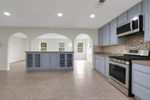 This spacious kitchen features modern stainless steel appliances, light teal cabinetry, and a stylish mosaic tile backsplash. The open layout includes a central island with shelving and arches leading to adjacent rooms, creating a bright and welcoming atmosphere.
