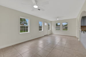 Bright, spacious living area with large windows, tiled floors, and ceiling fans. Features neutral tones and an open layout, leading to the kitchen with arched openings and access to the backyard