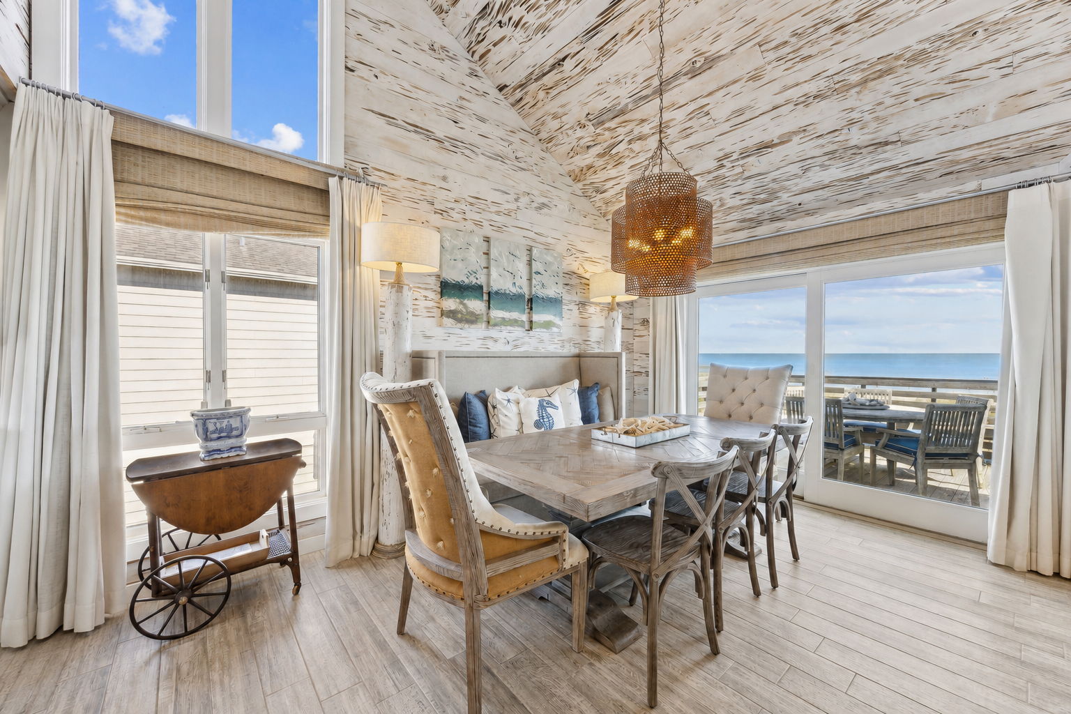 The dining area continues the coastal story with soaring ceilings, wood-grain tile flooring, and ocean views beyond the glass...