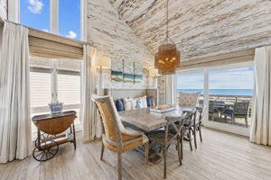 The dining area continues the coastal story with soaring ceilings, wood-grain tile flooring, and ocean views beyond the glass...